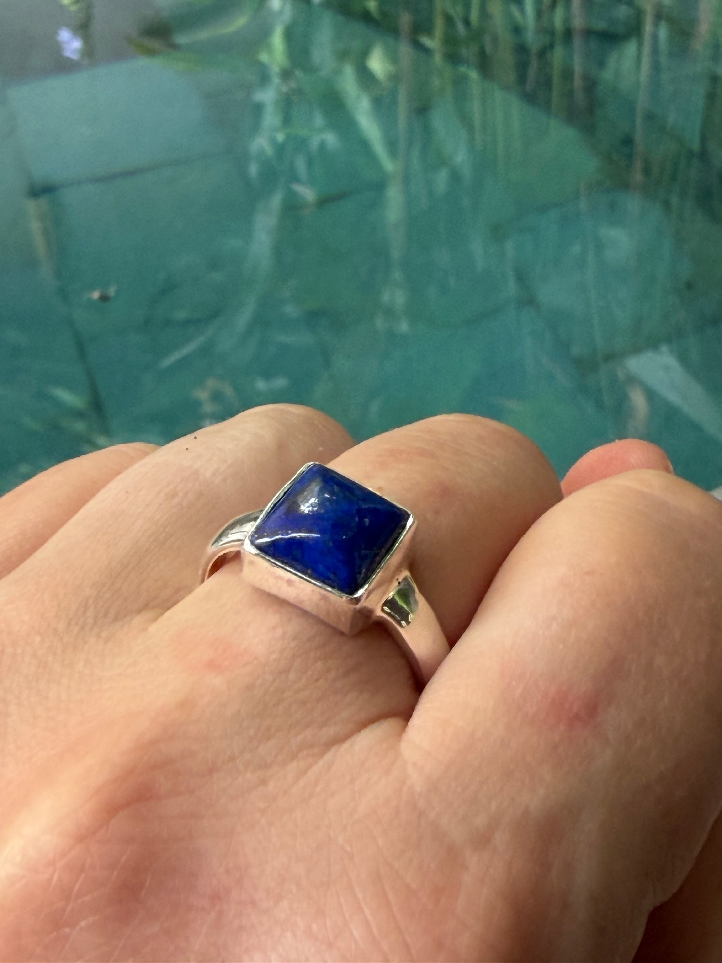 Hand wearing a ring with a blue Lapis Lazuli gemstone against a blurred green background